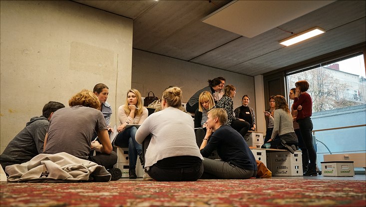 Zwei Gruppen von Teilnehmenden sitzen nebeneinander auf dem Boden oder auf leeren Getränkekisten und diskutieren über das Thema ihrer Session.