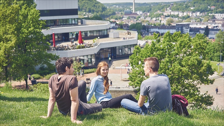 Drei Studierende sitzen auf einem Hügel auf der Wiese, im Hintergrund ist ein Universitätsgebäude zu sehen