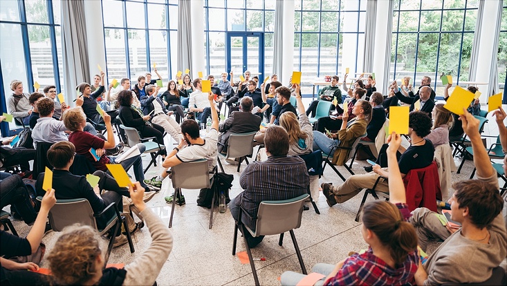 Gruppe Menschen in einem gläsernen Gebäude, sitzend von hinten fotografiert, einige heben den die Hand mit einer Abstimmungskarte