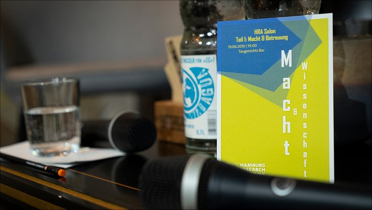 Auf einem Tisch liegen zwei Mikrofone und ein Flyer des HRA Salons zum Thema "Macht und Wissenschaft".