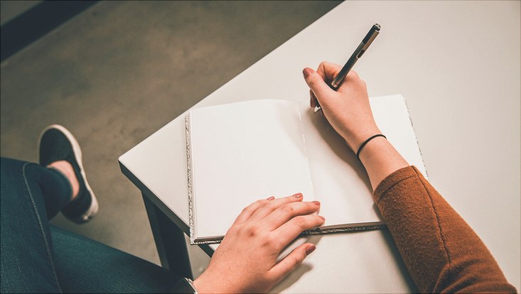 Eine Person sitzt an einem Tisch. AUf dem Tisch liegt ein Heft in dem die Person gerade etwas schreiben will. Die Seiten sind leer. Die Perspektive ist von oben.
