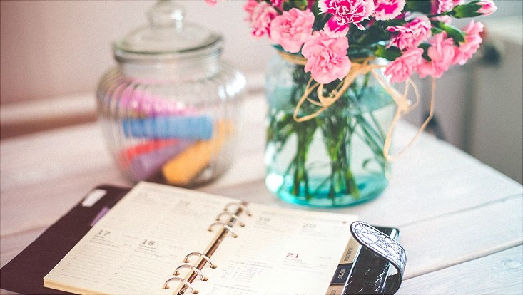 Ein aufgeschlagener Kalender liegt auf einem Tisch, im Hintergrund Blumen