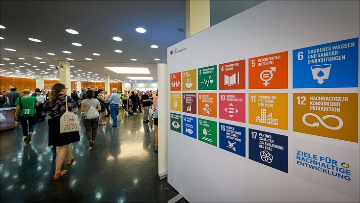 Konferenz. Auf der linken Seite laufen Menschen. Auf der rechten Seite ist ein Plakt mit den 17 Sustainable Development Goals.