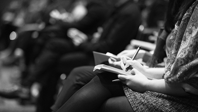 Das Bild zeigt eine sitzende Frau in einer Reihe von Tagungsteilnehmenden, die auf ihren Knien ein Notizbuch liegen hat, in dem sie Gehörtes mitschreibt.