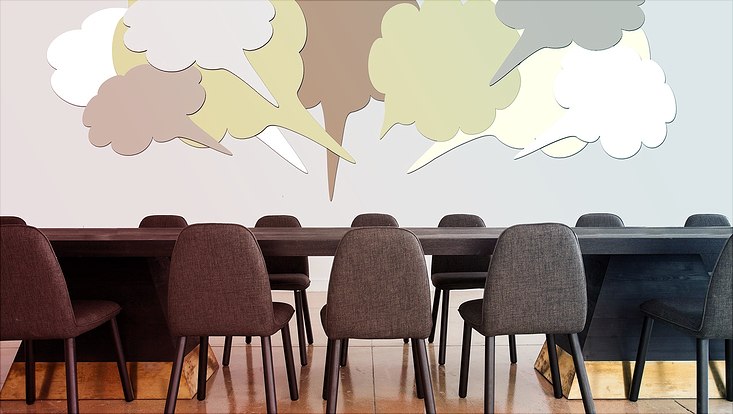 Stühle vor einem Tisch über dem grafisch dargestellte Sprechblasen schweben