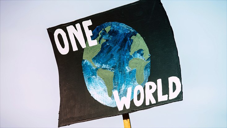 Auf einem Schild ist der Schriftzug "One World" sowie ein gemalter Erdball zu sehen.